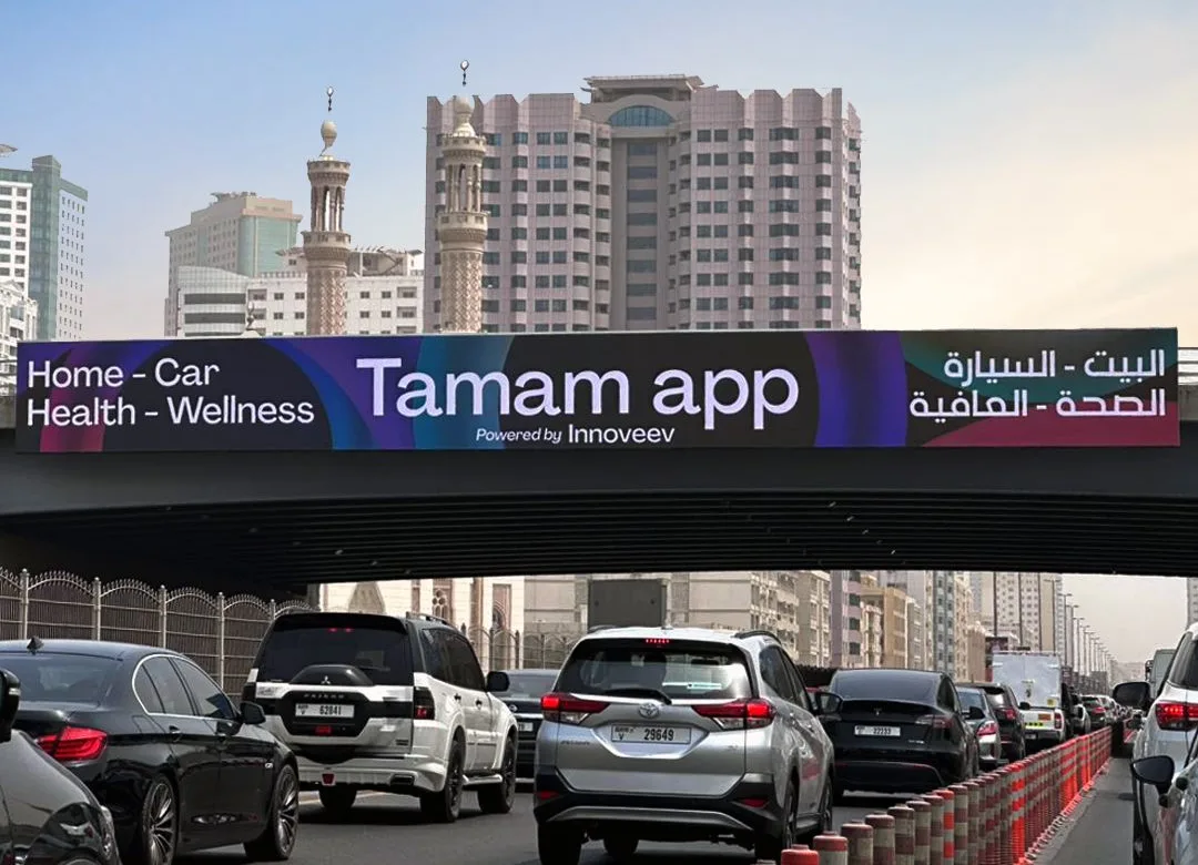 A Tamam app bridge banner spanning a busy road in Sharjah, with the slogan Home, Car, Health, Wellness in English and Arabic above lanes of traffic