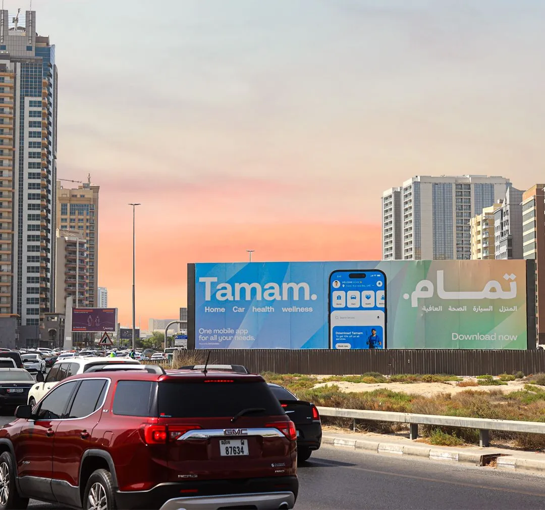 A large Tamam billboard beside a road in Sharjah at sunset, showing the app interface on a phone with the words Home, Car, health, wellness in English and the Arabic equivalent